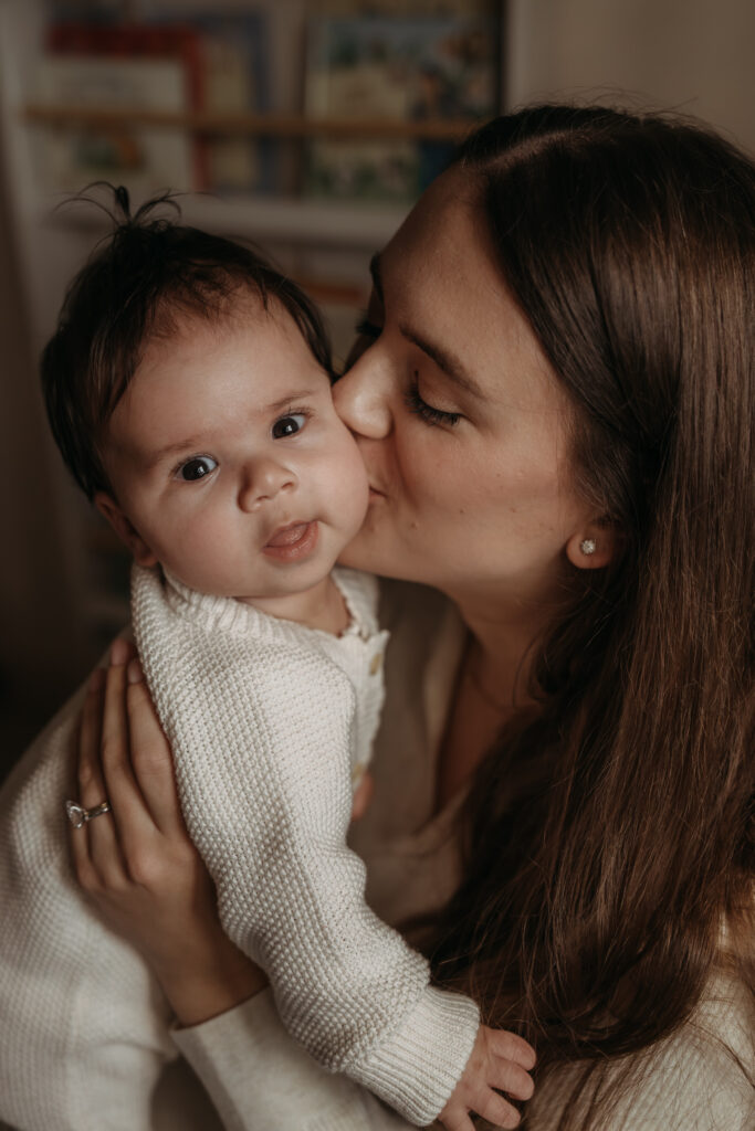 A close-up image of a Mum kissing the baby on a cheek and the baby stairs at the camera during a photo shoot at home