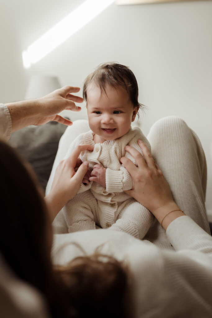 Four month old baby smiling at Mum whilst Dad reaches towards her during a family photo shoot at Home Hampshire