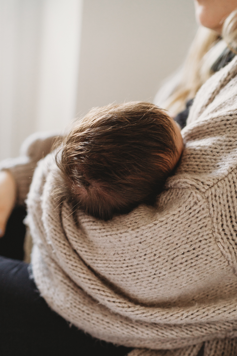 close up photo of a Newborn baby laying in his mothers arms with a full head of dark brown hair, she is wearing a beige knitted cardigan