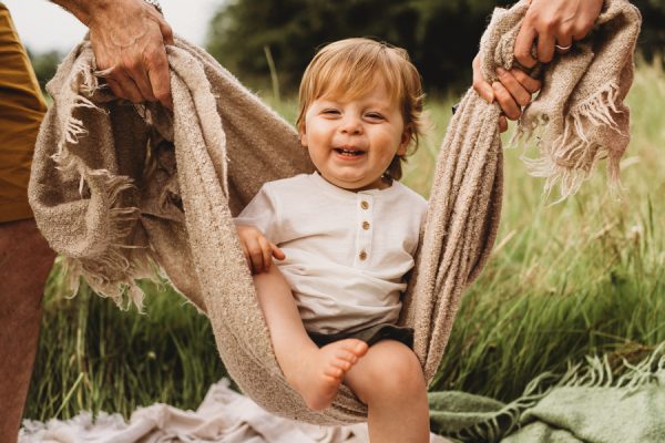 little boy swinging in a backlit held by her parents in the long grass meadow