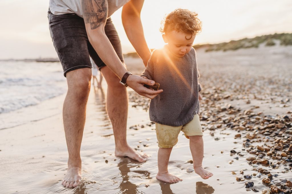 father and son holding hands on the beach with a sun flare bleeding through them 