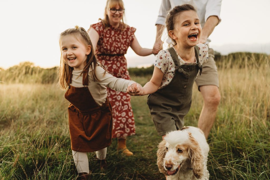 family running through the meadow with their dog chasing them whilst the sunsets behind them 