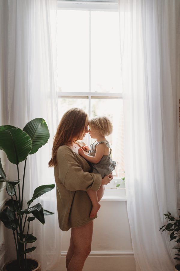 pregnant mama and her daughter balanced on her tummy standing in front of the window with sunlight streaming in