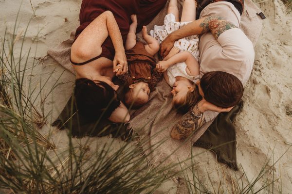 Family lying down cuddling on a blanket in the sand dunes nestled between the grasses