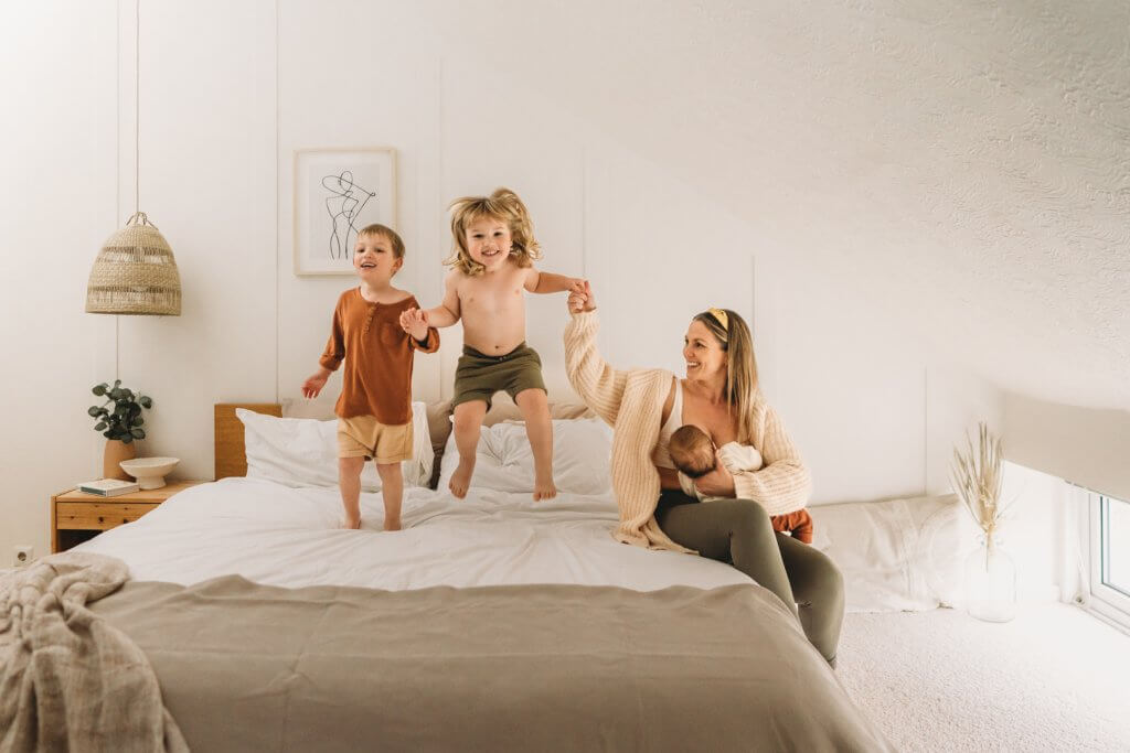photo of mum and sons playing/ jumping on the bed at home whilst new baby feeds