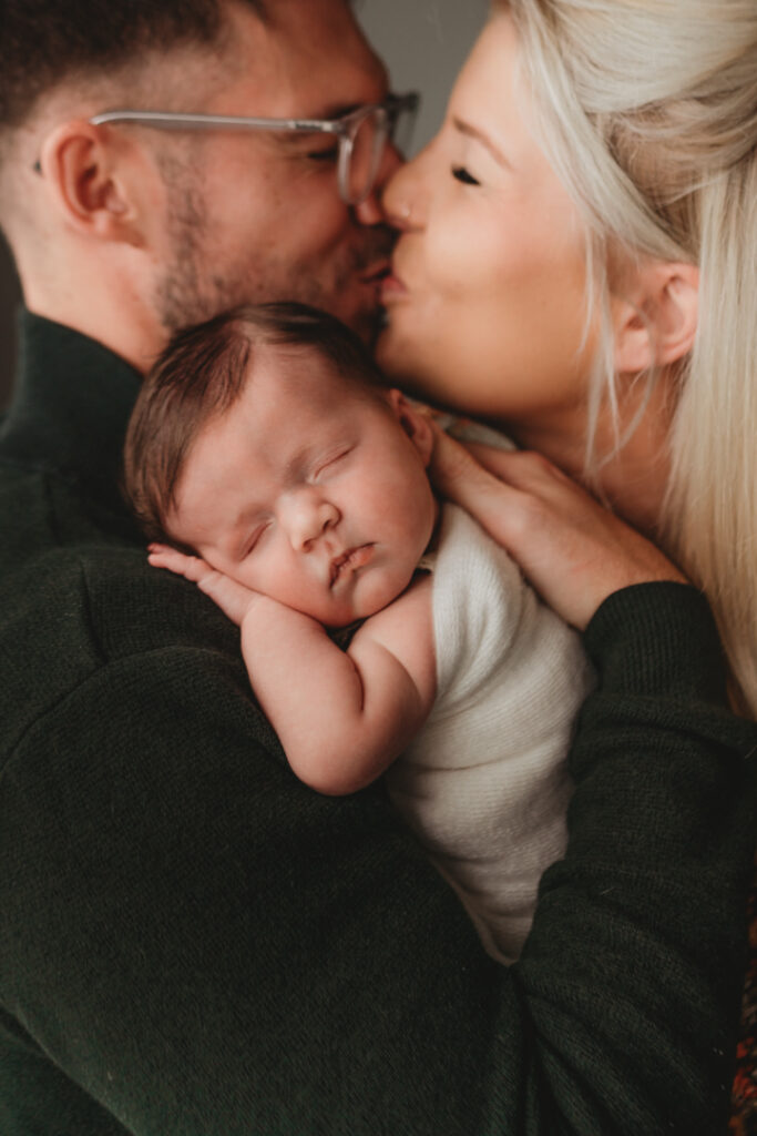 newborn sleeping on dad's shoulder mum and dad kiss behind her